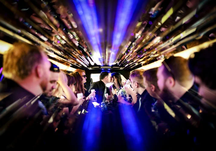 Newlyweds share a kiss in the limo while wedding party ookin at them