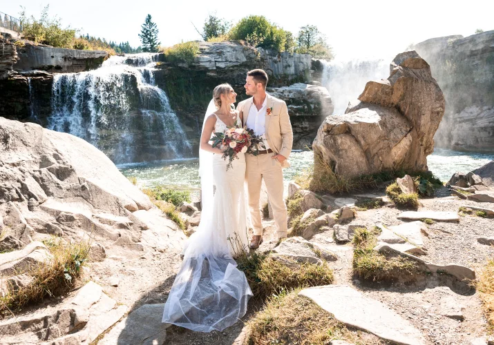 Bride an groom stand next other against stunning waterfall in Waterton.