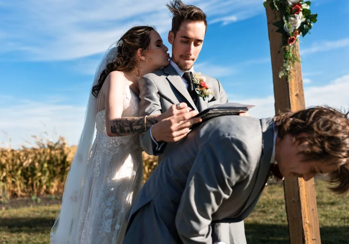 Groom sgh the wedding papers on the best-men back while the bride kisses him on the cheek.