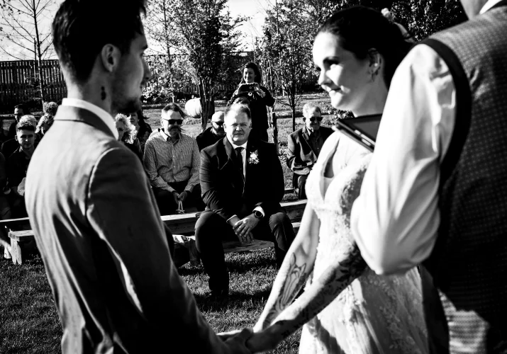 Black and White Wedding Photography by Calgary Wedding Photographers of the father of the bride looking an the newlyweds holding hands during the wedding ceremony.