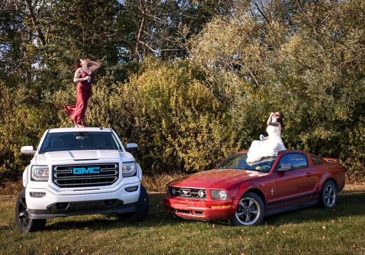 Bride and bridesmaid sitting on their cars and drink a beer.