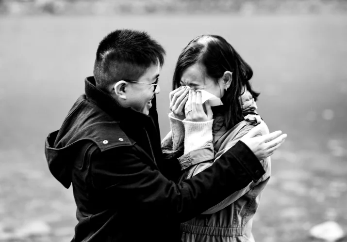Lady cries and whiles her eyes when her fiancee proposed. He hugs her.