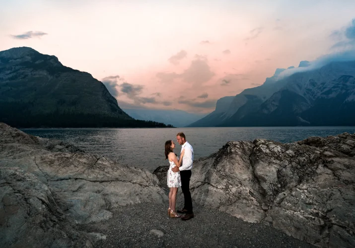 Engaged couple facing each other at Minnewanka Lake during colorefu sunrise