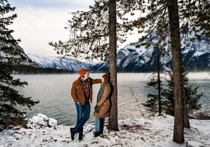 Lady leans over a tree at Minnewanka Lake while her fiancee look at her with love.
