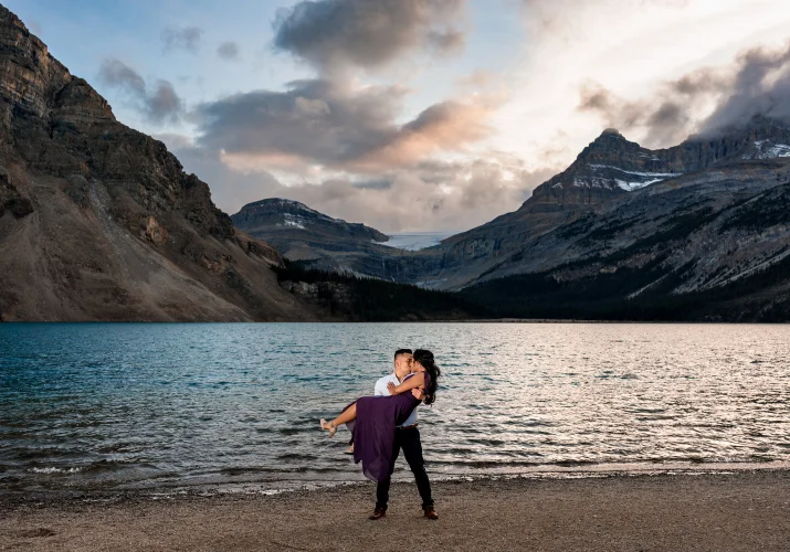 Man lifts his fiancee up at Bow Lake against huge mountains.