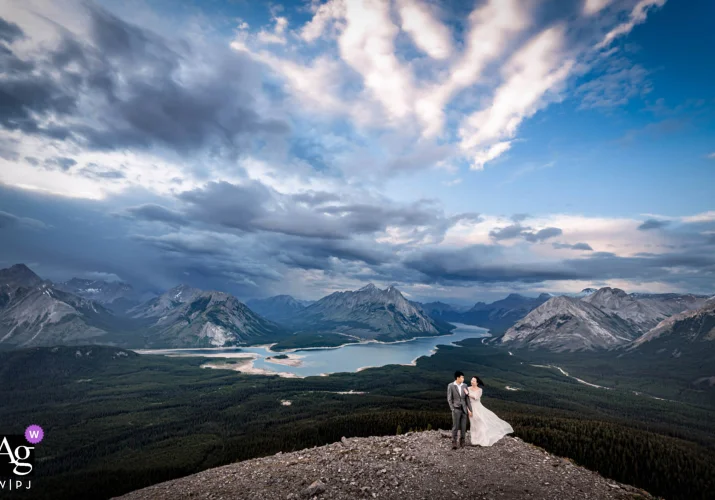 Bride and groom standing next to each other at the top of the mountain. In the background a blue lake and huge Canadian Rockies.