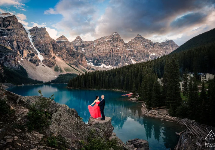 Man and a lady in a red dress stand on the rock at Moraine Lake. On the background Rocky Mountains and blue lake.
