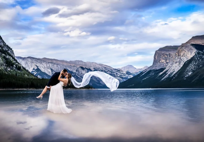 Creative photograph of newly weds dancing at the lake. Bride's veil flow with the air. They are surrounded by the Canadian Rockies at blue Minnewanka Lake . Photo taken by the best wedding photographer 4Eyes Photography.