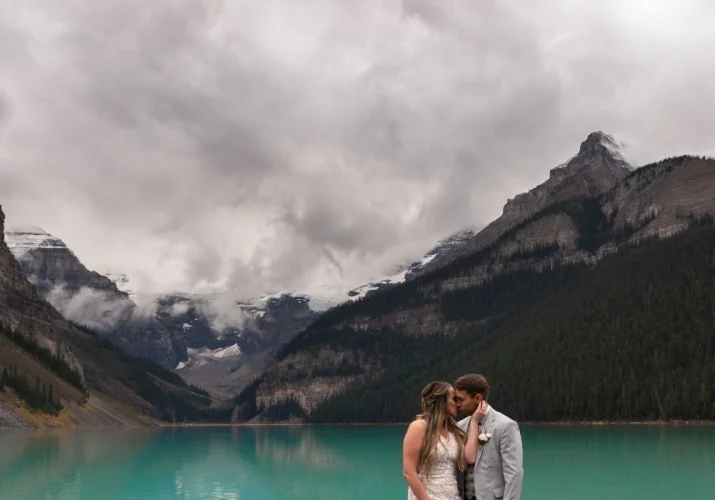 Beautiful bride kissing her new husband at Lake Louise which is surrounded by the mountains