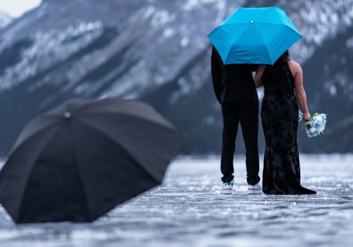 Newly engaged couple walks under a blue umbrella. They look at the beautiful mountain in Banff.