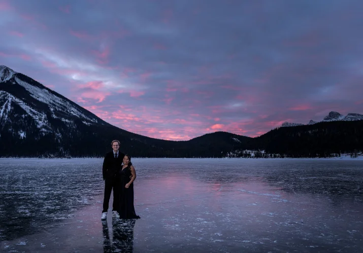 Stunning sunrise at Minnewanka Lake in Banff. Couple in love stand at the frozen lake an look at the photographer during photo session.