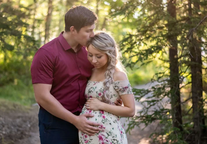 Maternity photo session in Banff. A man touches his pregnant wife's belly and kisses her forehead.