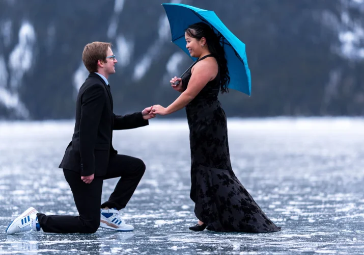 Romantic proposal on one knee in Banff. Future bride cover herself under a blue umbrella.