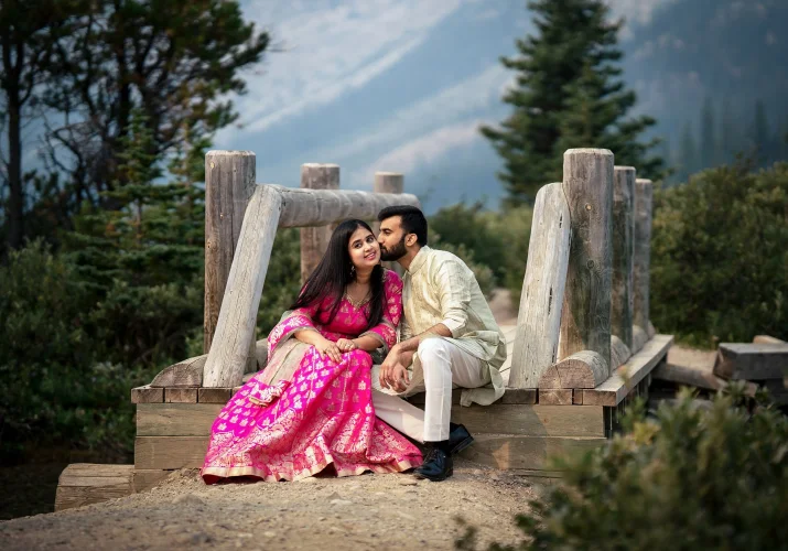 Indian couple wears traditional outfit. Man is kissing his fiancee on her cheek. They are sinning at the wooden bridge at Bow Lake.