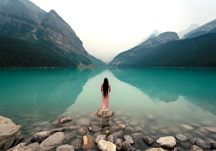 Lady in a pink dress standing at the rock at Lake Louise. She is looking at stunning Canadian Rockies.