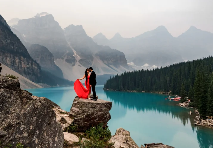 Engagement photo session at Moraine Lake captured by Banff Wedding Photographers. Beautiful coupe standing on the rock. Woman is wearing a red, long dress and her fiancee is wearing a black suit.