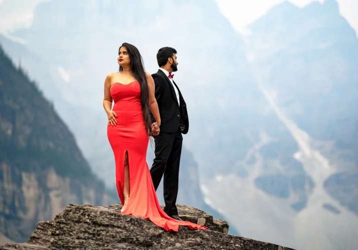 Lady in red dress look far away and her fiancee in suite look opposite direction. They are surrounded by the massive mountains in Banff.