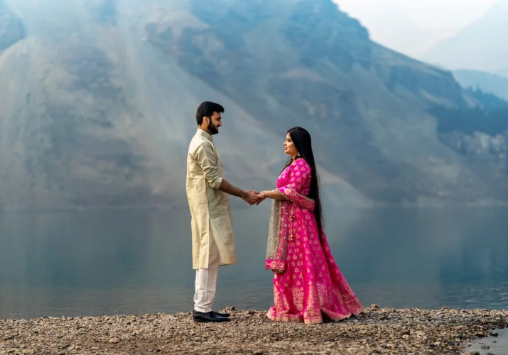 Indian couple wear traditional outfit. She wears a pink dress and he wears gold traditional outfit. They hold their hands and look at each other at Bow Lake in Banff