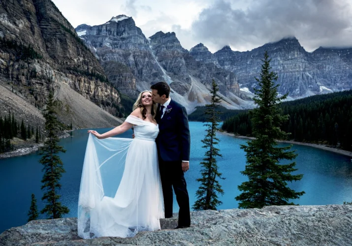 Newlyweds standing at the giant rock at Moraine Lake. They are surrounded by the Canadian Rockies. She holds her dress and he kisses her cheek.