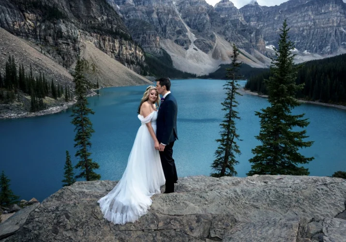 Groom kissing the bride at her forehead. They holds their hands at Moraine Lake in Banff.
