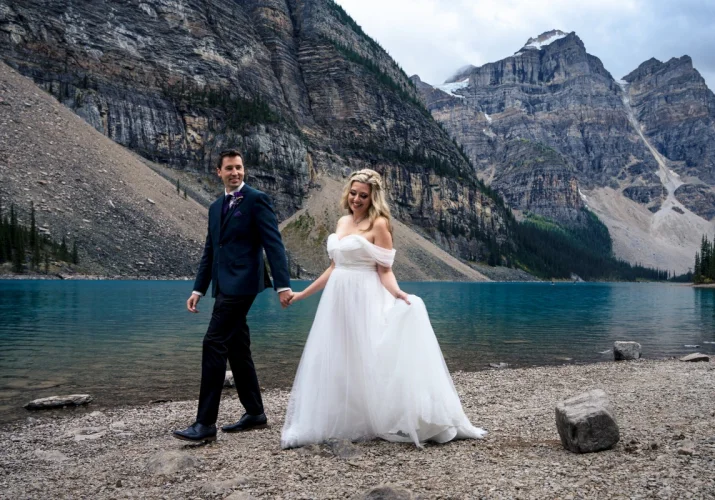 Bride and groom walk at Moraine Lake holding their hands. Groom is looking at his gorgeous wife.