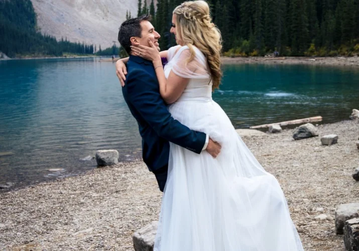 Groom lifts his wife. She puts her hand on his cheek. They are at the Moraine Lake.