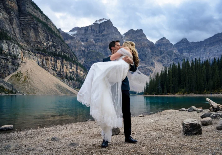 Groom holds the bride on his hands. They are kissing surrounded by the Canadian Rockies in Banff.