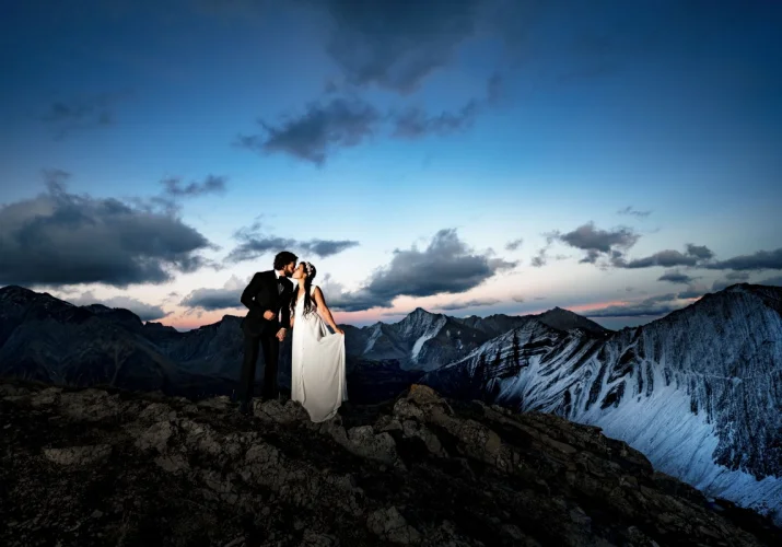 Banff Wedding Adventure Photo Session at the top of the mountain. Newlyweds hold their hands and kiss each other. Mountains are covered with snow and sun goes down.