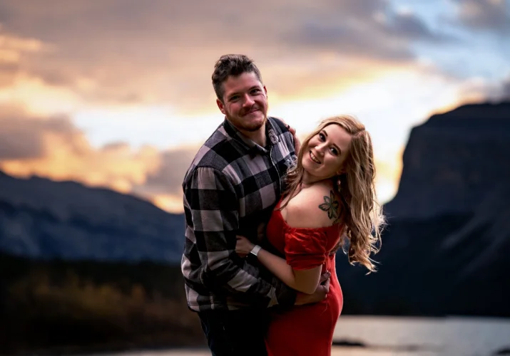 engaged couple look at the camera and smile during sunrise at Minnewanka Lake in Banff