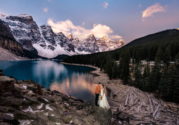 Newlyweds adore pink sunrise at Moraine Lake in Banff. They stand at the rock and look at the blue lake.