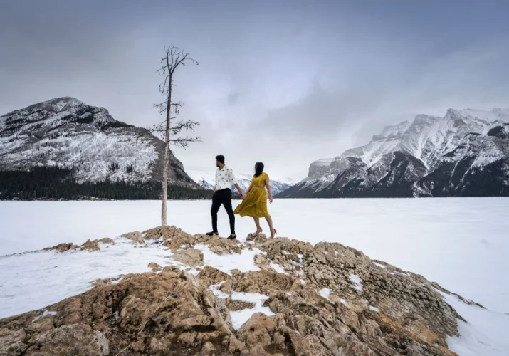 Engaged couple standing at the giant rock by frozen Minnewanka Lake. They look forward them and admire mountains.