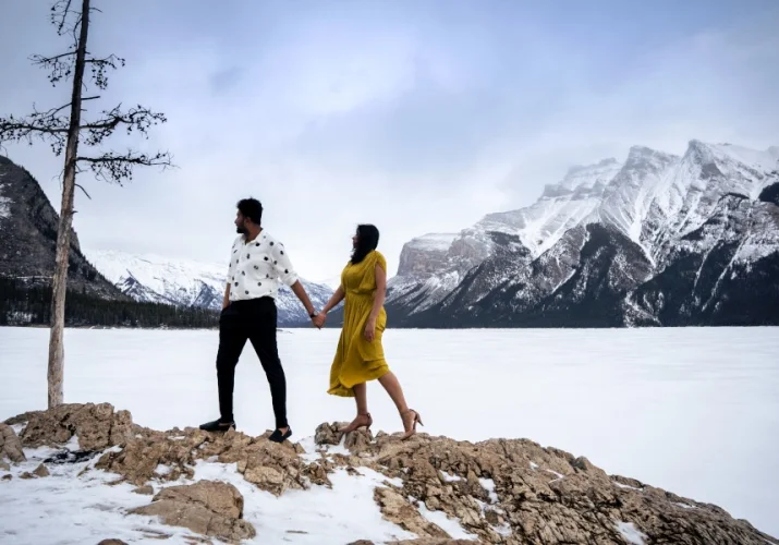 Man and woman stand at the edge of the frozen Minnewanka Lake. They are surrounded by Canadian Rocky Mountains.