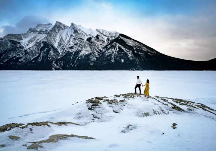 Incredible engagement photography taken by Calgary Wedding Photographer at Minnewanka Lake in wintertime. Couple standing at the giant rock and admire the beauty of the mountains.