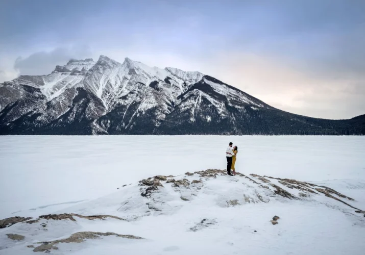 Engagement Photo Session at Minnewanka Lake in winter time. Lake and mountains are covered with white snow. Dressed, engaged couple hug each other.