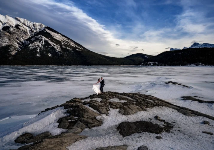 After wedding session at Minnewanka Lake with the best photographer Eliza Slobodzian. Couple stands at the frozen lake. They are surrounded by the Canadian Rockies.