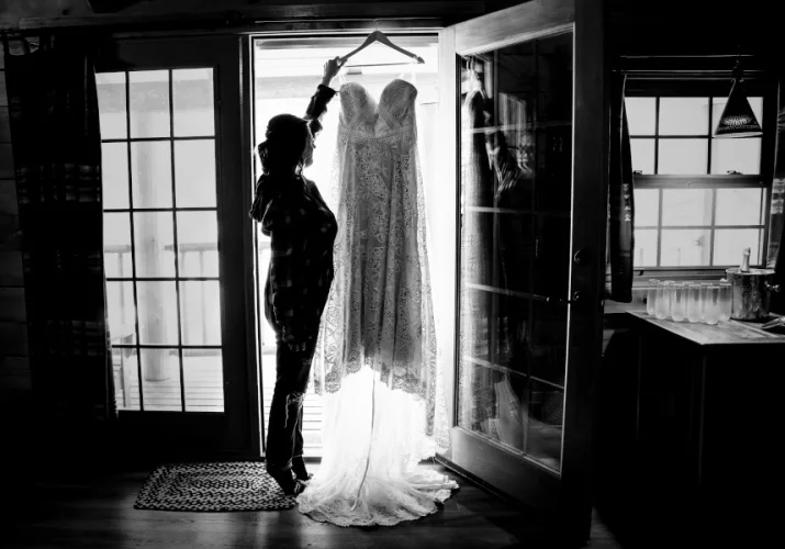 Bride reaches for her white wedding dress which hangs on the large window.