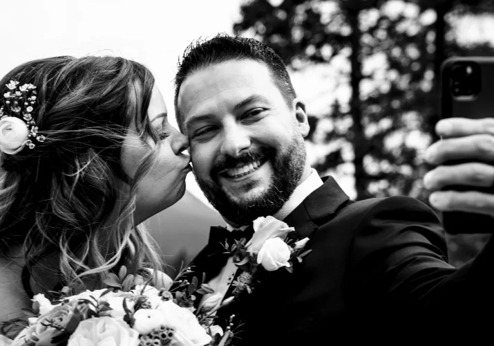 Bride kisses cheek of her husband. The broom takes selfie of them.