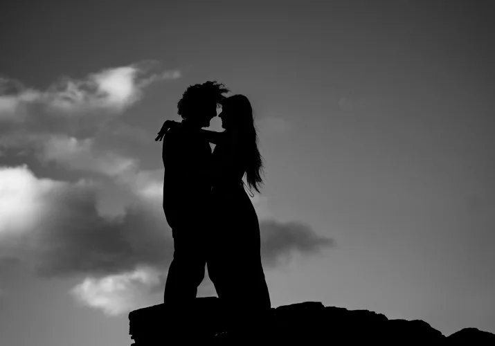 silhouette photograph of engaged couple in Black and white. They hug each other on the rock. In the background is sky.