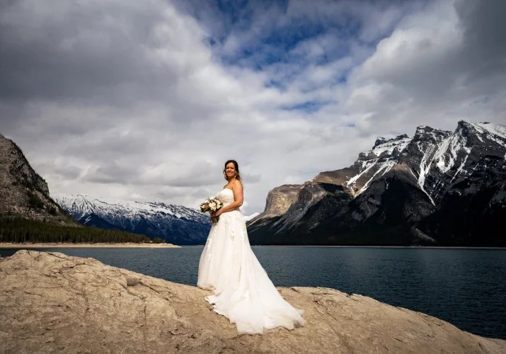 Bride's portrait. She stands on the big rock by the Minnewanka Lake. She holds bouquet and smiles.