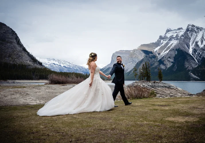 Bride and groom walk down the Minnewanka Lake . They hold their hands and look at each other.