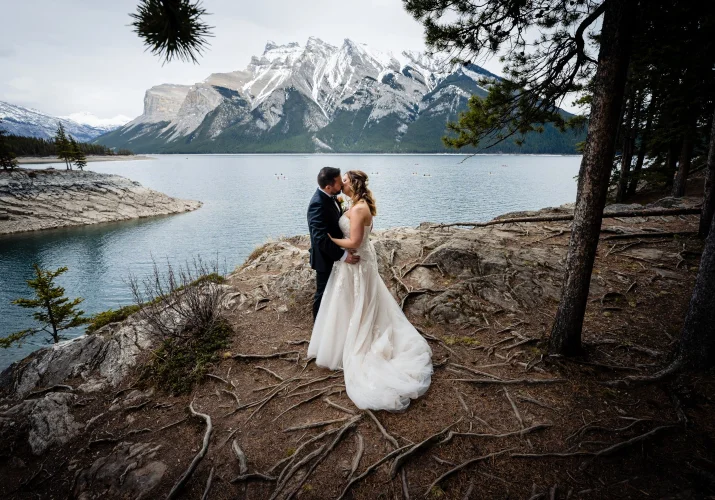 Newlyweds kiss during their wedding photo session at Minnewanka Lake. On the background big mountain covered with snow.