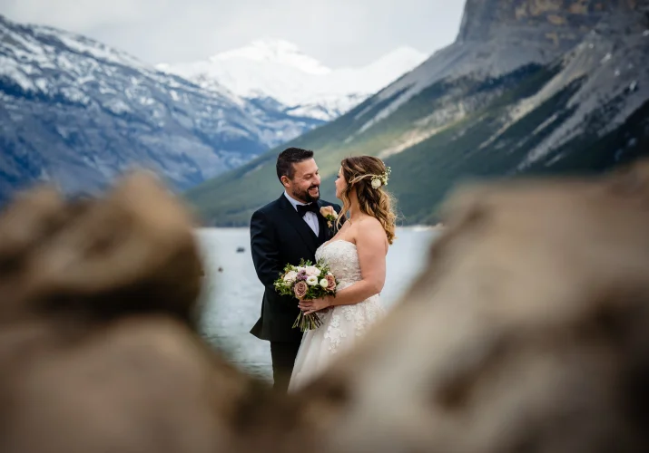bride and groom look at each other and hug. Stunning mountains and blue Minnewanka Lake on the background. They ate kept in the frame of the rocks.