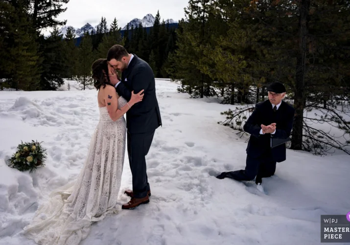 First kiss of bride and groom during Banff elopement. Officiant clapping hands fall down into deep snow.