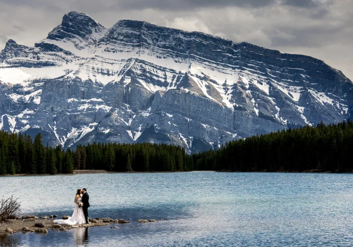 Newlyweds hug during wedding photo session captured by photographer Eliza Slobodzian. Couple is surrounded by a giant mountain and Two Jack Lakes in Banff.