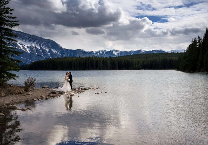 Newlyweds standing at the edge of Two Jack Lakes in Banff and hug. Amazing Canadian Rockies on the background.