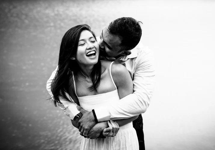Man whisper a secret to his fiancee ear. She laughs. Black and white photography taken by best Banff Wedding Photographer.