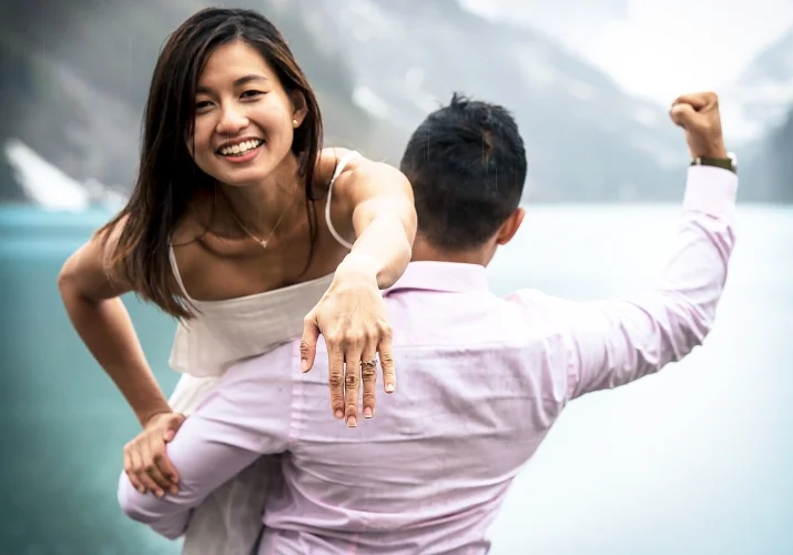 Man carry his fiancee on his shoulder while she laugh and shows her hand with an engagement ring.