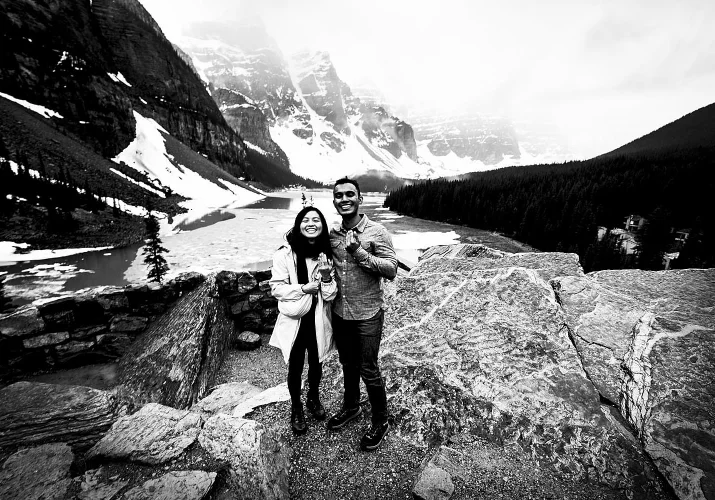 A couple shows their engagement rings. They smile at Moraine Lake.