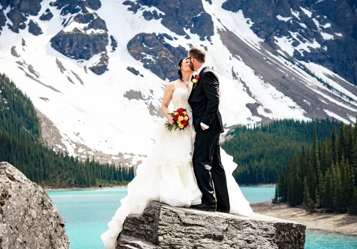 Newlyweds share a kiss at Moraine Lake in Banff. They are surrounded by snowy mountains.