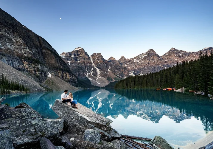 Couple in love sits at the huge rock by the turquoise Moraine Lake against breathtaking Canadian Rocky Mountains.
